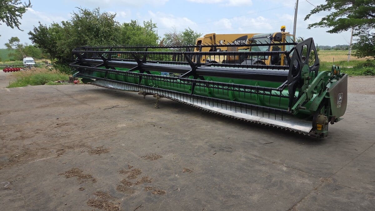 John Deere 930F flex grain header with its model badge, resting in our Albion, Iowa warehouse yard beside a telehandler