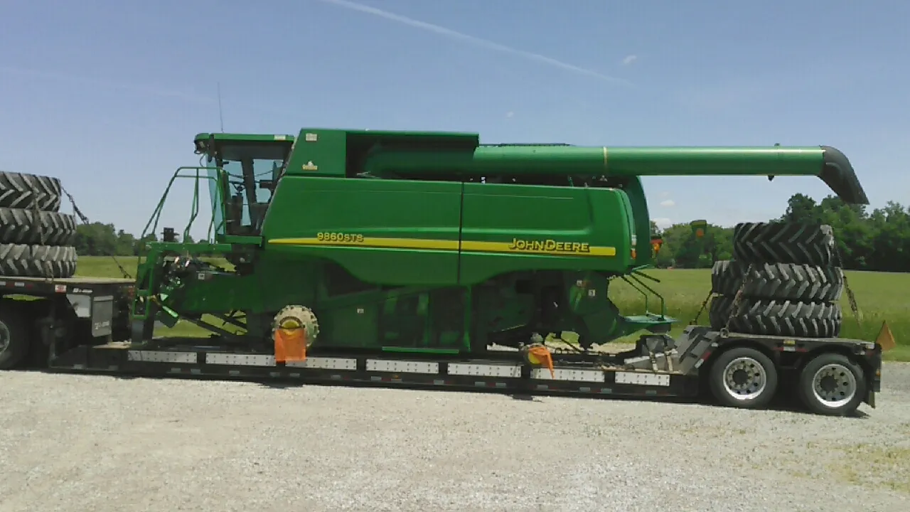 John Deere combine on a low trailer with large tires stacked at both ends of the deck, open fields behind