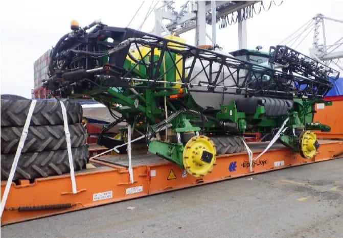 Sprayer with a folded boom on a flat rack at a container terminal, strapped tires at its side and cranes behind