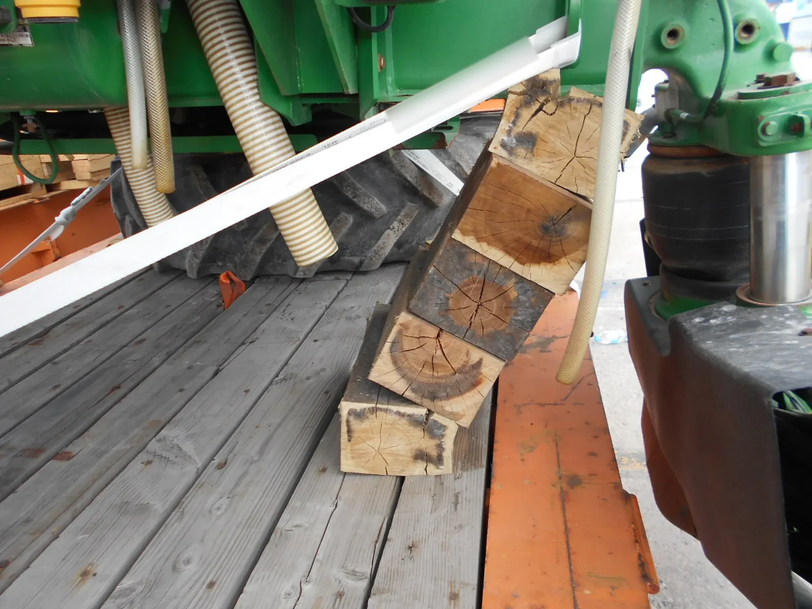 Close-up of timber blocks tilted on a wooden flat rack deck beside a tire, with a white lashing strap across the frame