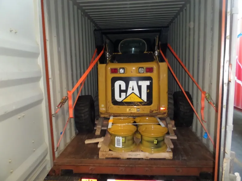 Погрузчик Caterpillar, закреплённый ремнями в контейнере, перед ним колёсные диски на поддоне