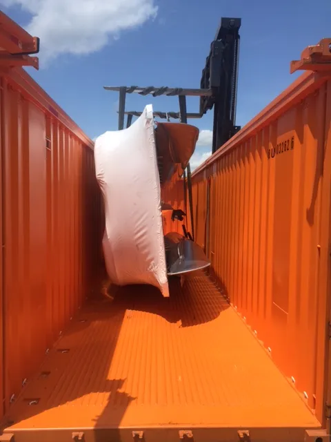 A boat wrapped in protective film inside an orange open-top container, with a forklift mast above