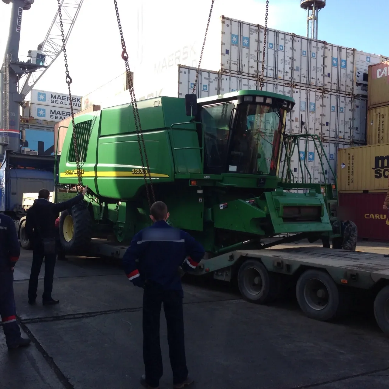 Combine hanging from chains above a lowboy trailer at a container port, with stacked containers and a crane behind