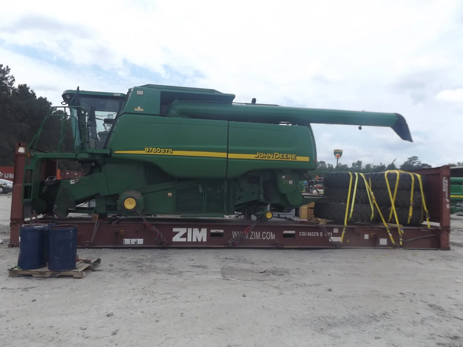 Combine on a flat rack with strapped tires at the rear and blue drums on a pallet at the front left
