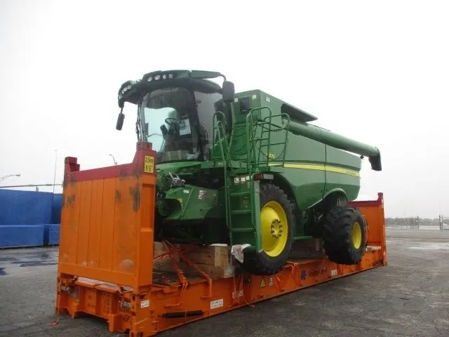A green John Deere combine with yellow wheels on an orange flat rack with its end walls up, in a paved yard