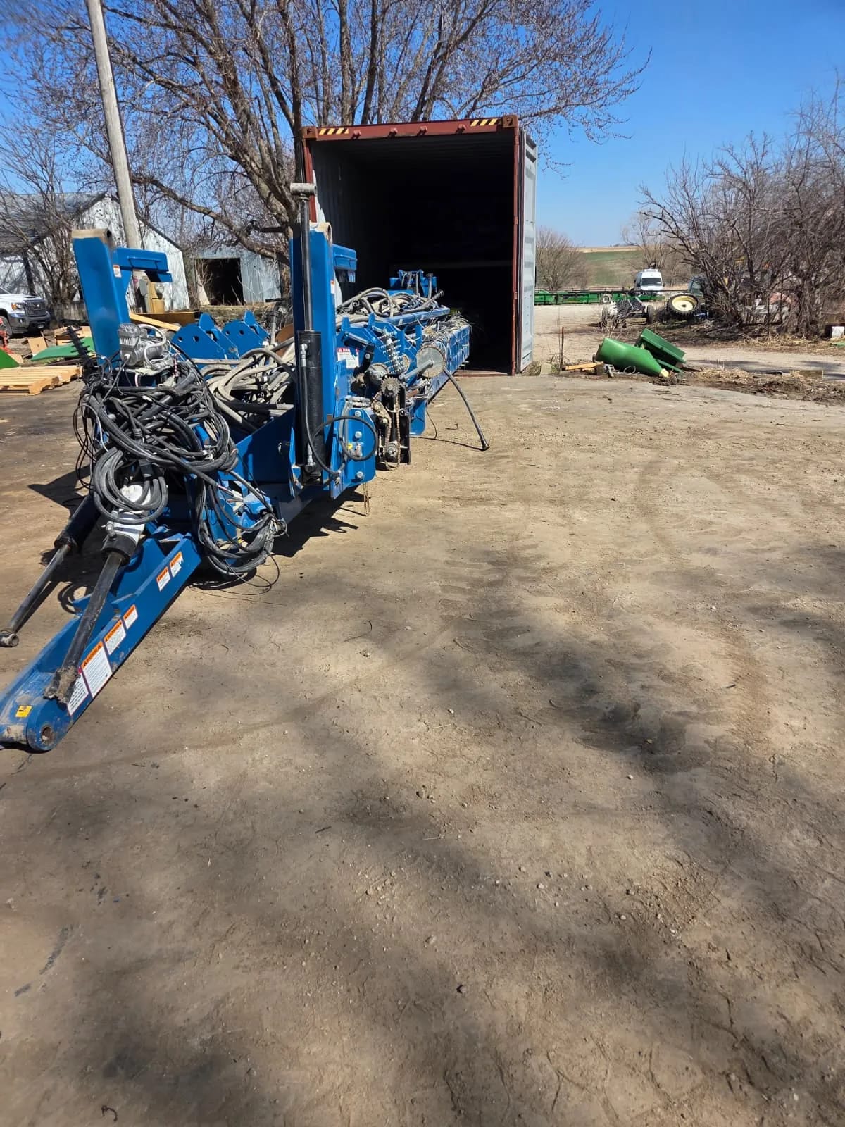 Folded planter frame with bundled hoses and wiring on a dirt yard beside an open shipping container