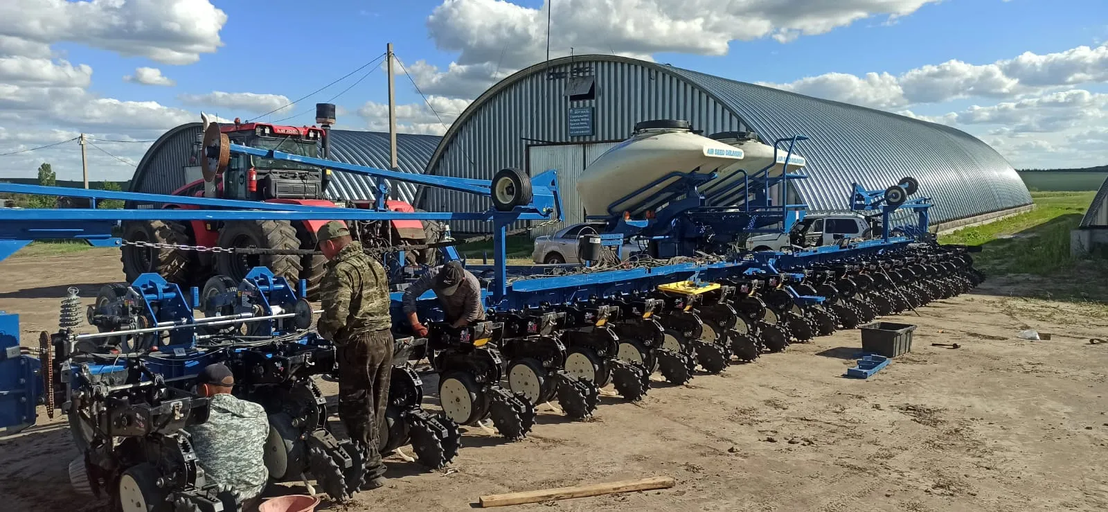 Planter with a blue frame laid out in a farm yard, people at its row units and a red tractor behind
