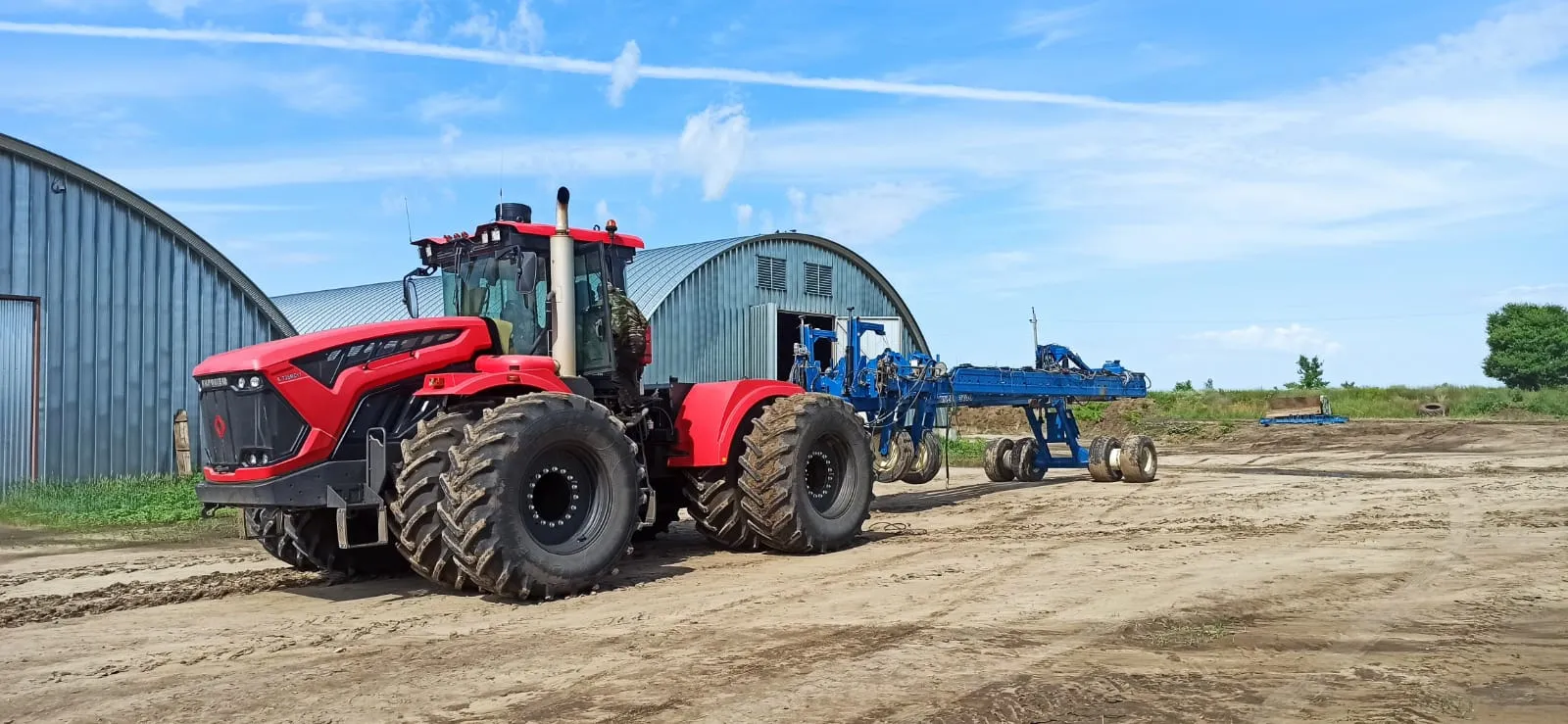 Tractor with a red cab and large dual wheels beside a blue planter frame in front of curved-roof hangars