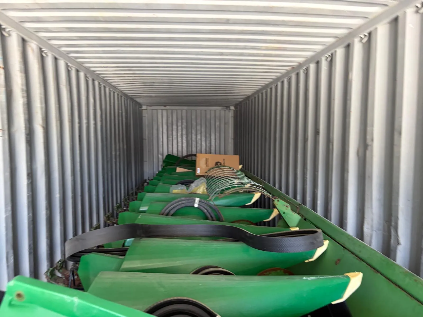 Corn header row units lined up along the side of a shipping container, with a belt looped over them