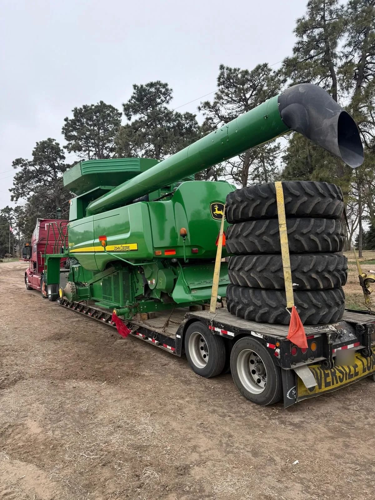 John Deere combine on a low trailer with four large wheels stacked and strapped at the rear of the deck