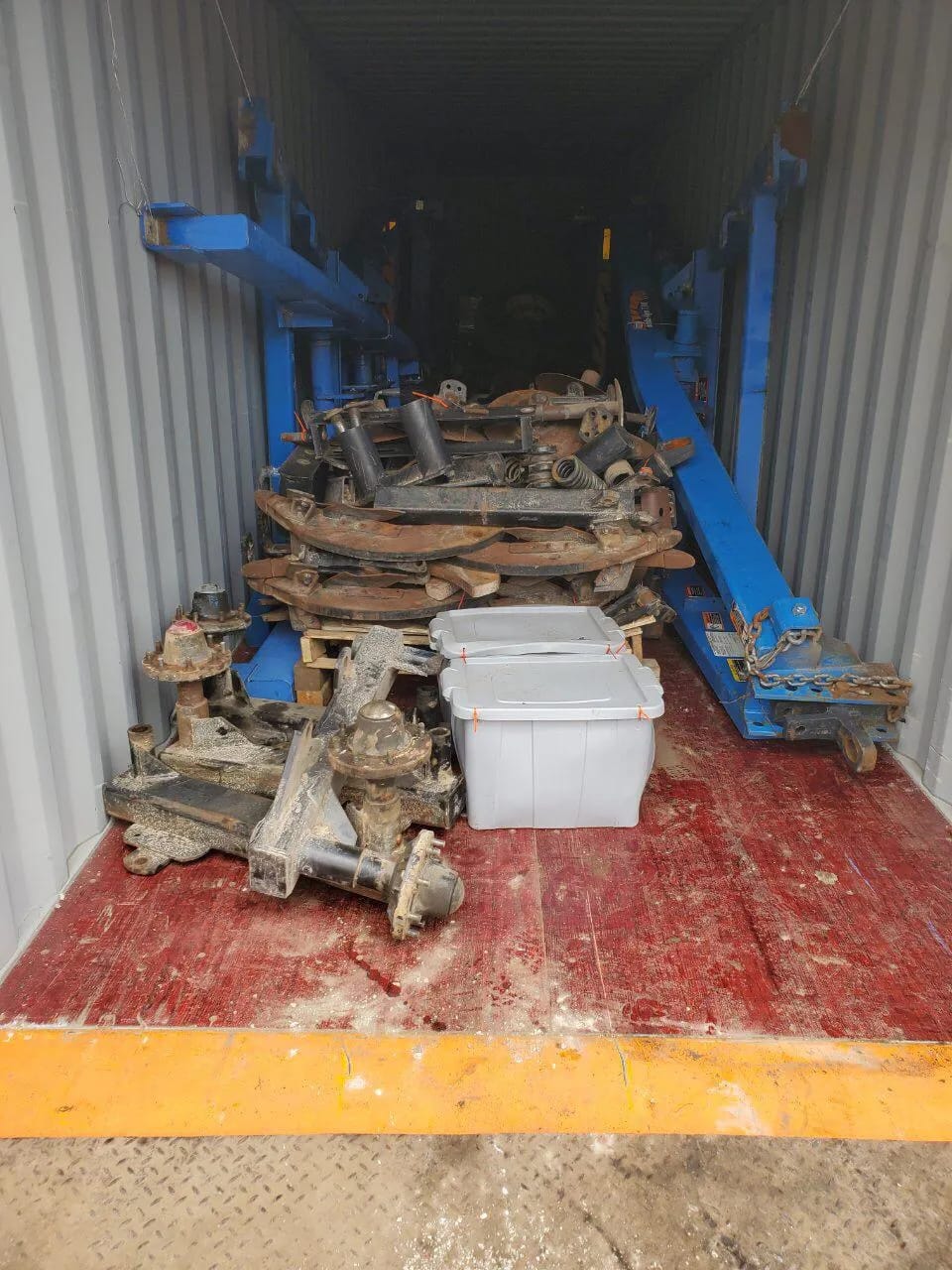 Implement frames in blue along the walls of a container, with rusty disassembled parts and plastic tubs on the floor