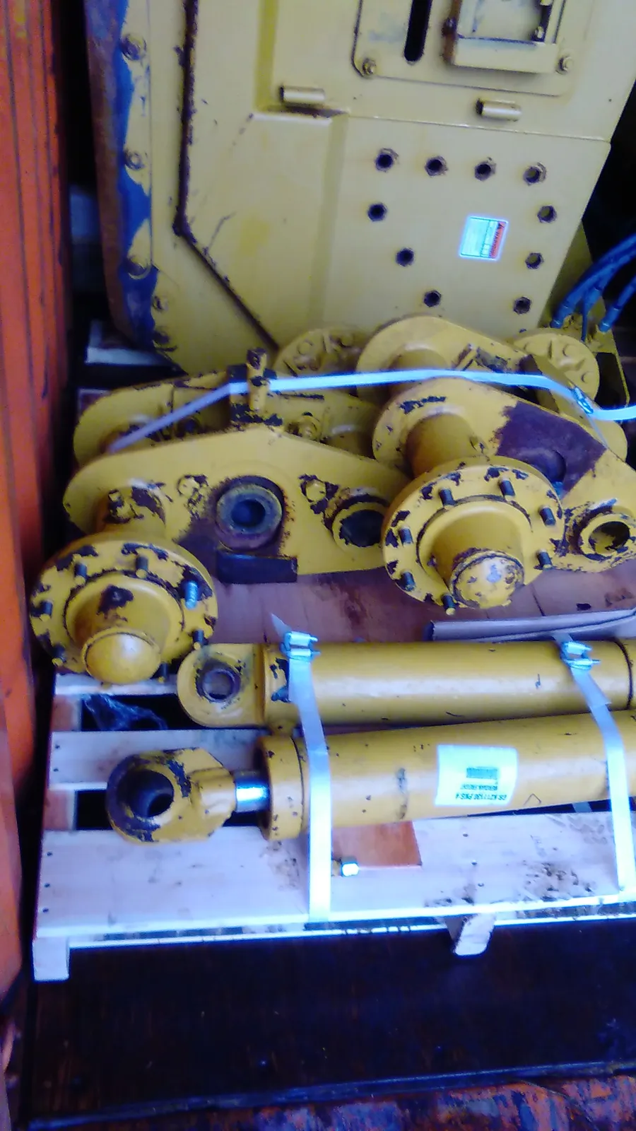 Parts strapped to a pallet: hydraulic cylinders below a yellow machine frame with rows of holes