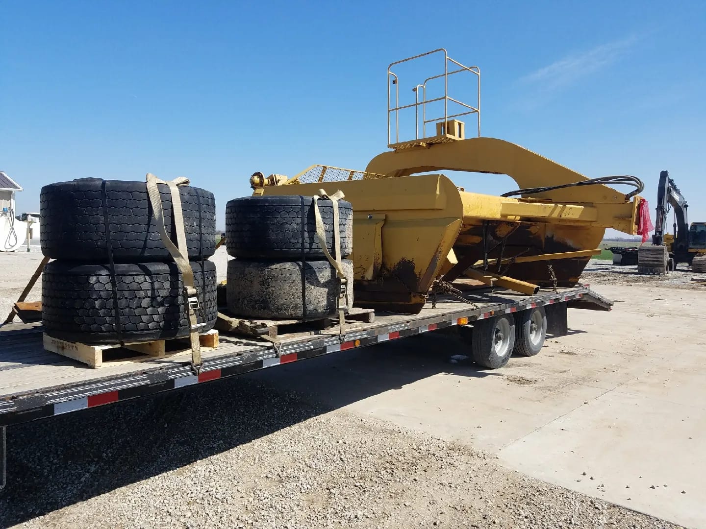 Leveler bowl and arched frame on a flatbed trailer with strapped tire stacks, an excavator behind