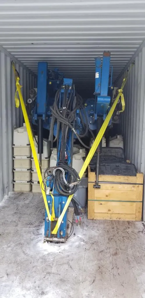 Planter frame held by yellow straps inside a container, with grey boxes at the left and a wooden crate at the right
