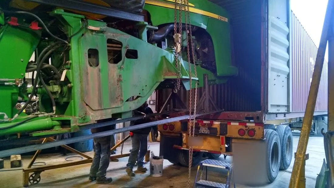 Disassembled tractor frame hanging from chains in a workshop beside a container on a truck trailer