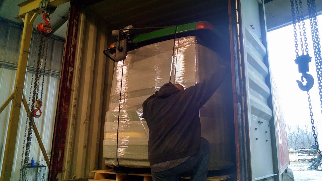 Machine section wrapped in film inside an open container as a worker reaches up, chain hoists hanging at both sides