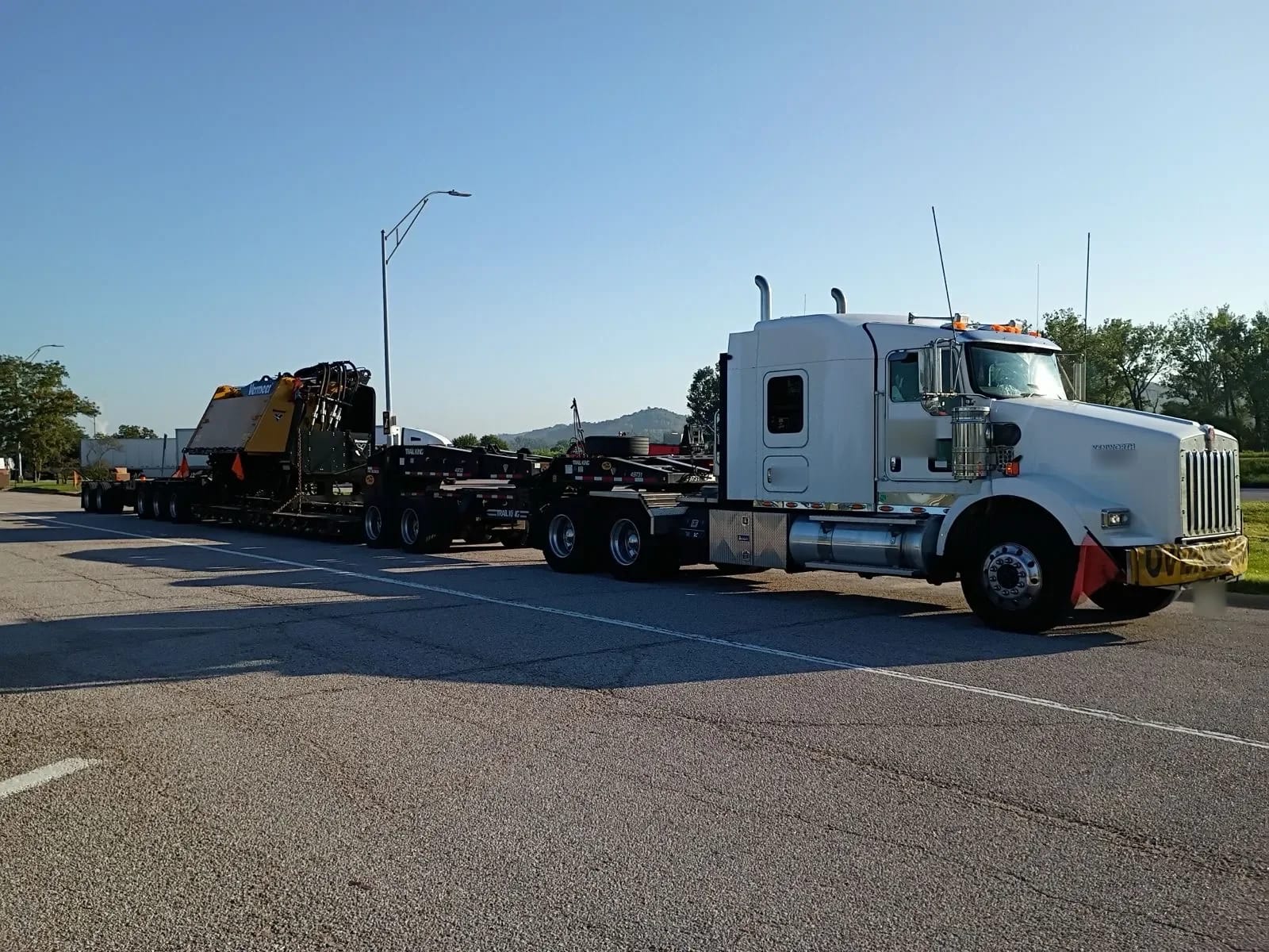 Long multi-axle lowboy trailer with a Vermeer attachment, pulled by a white truck on a paved road
