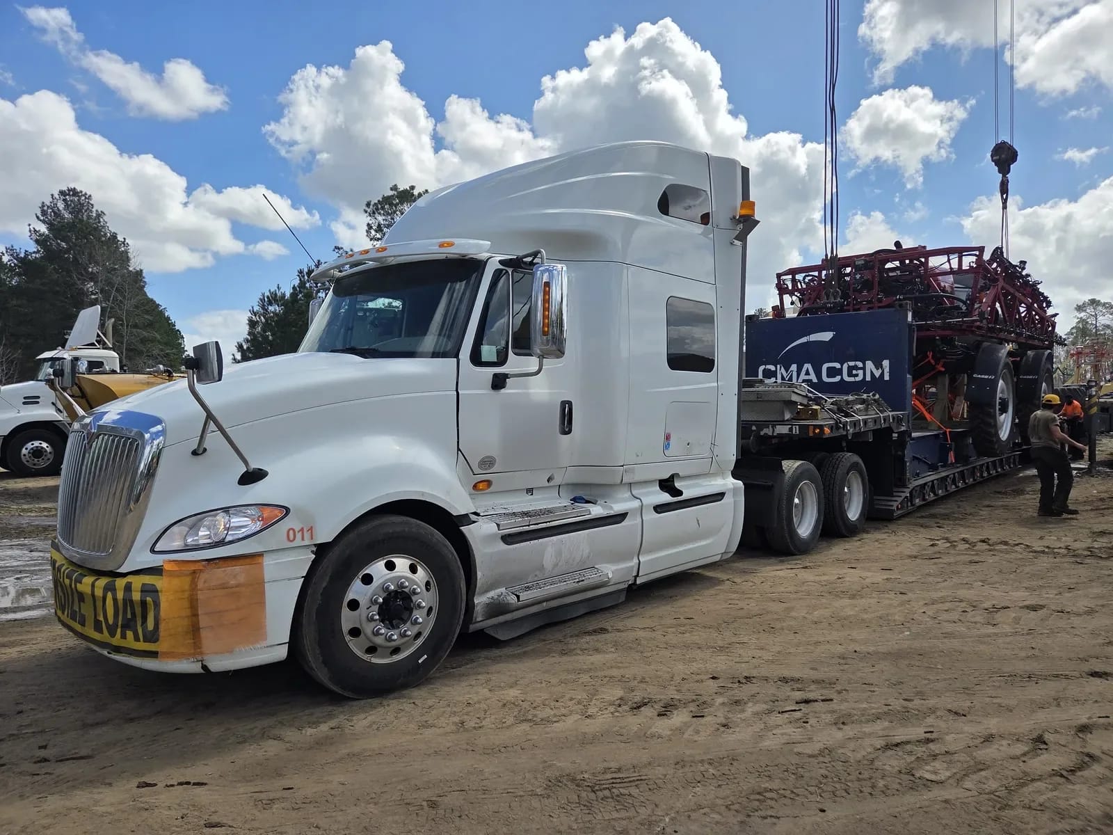 A truck with a lowboy trailer and a red sprayer being lifted by a crane at a dirt yard
