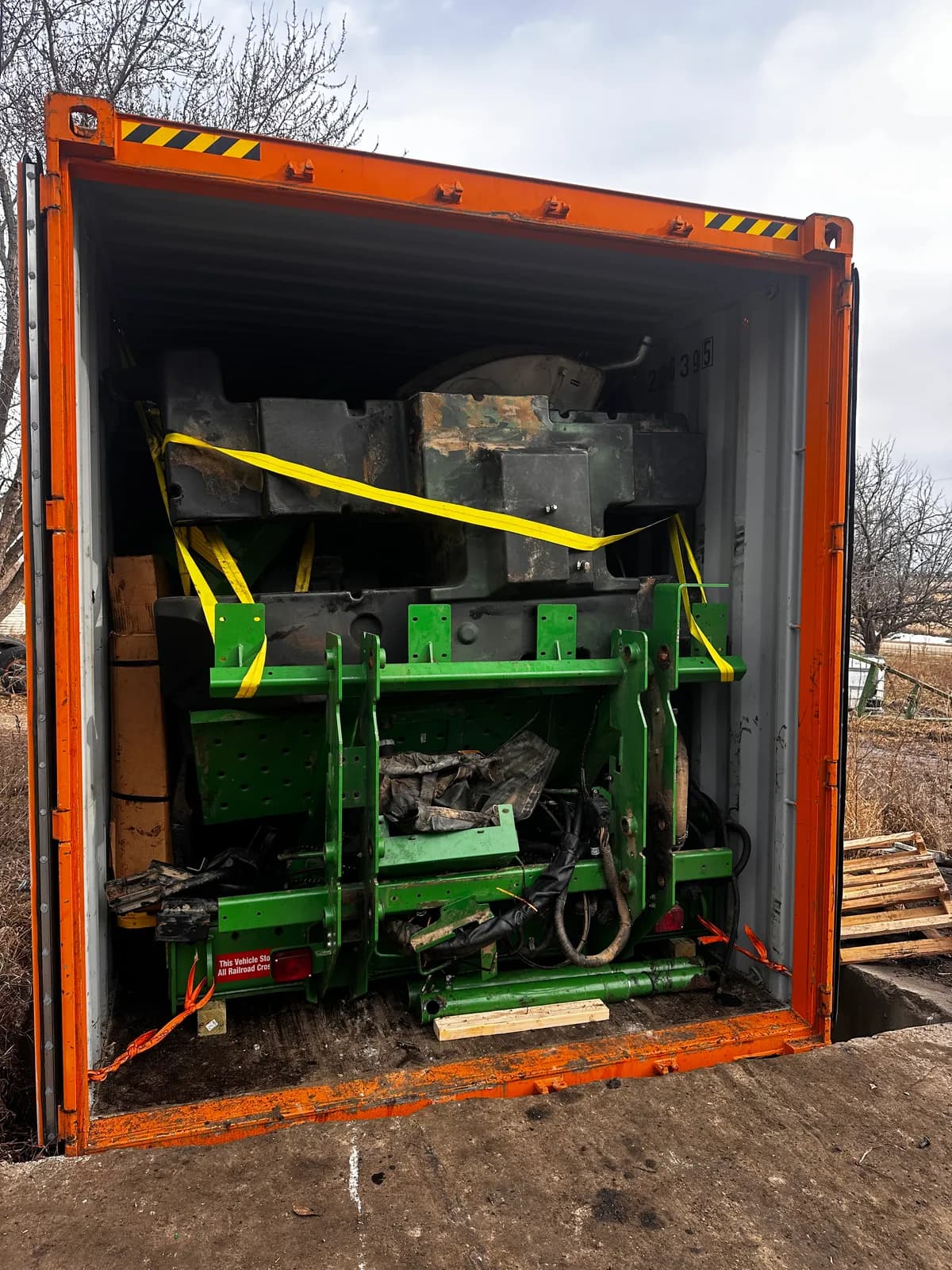 Dismantled sprayer chassis and heavy parts strapped inside an open container on wooden blocking
