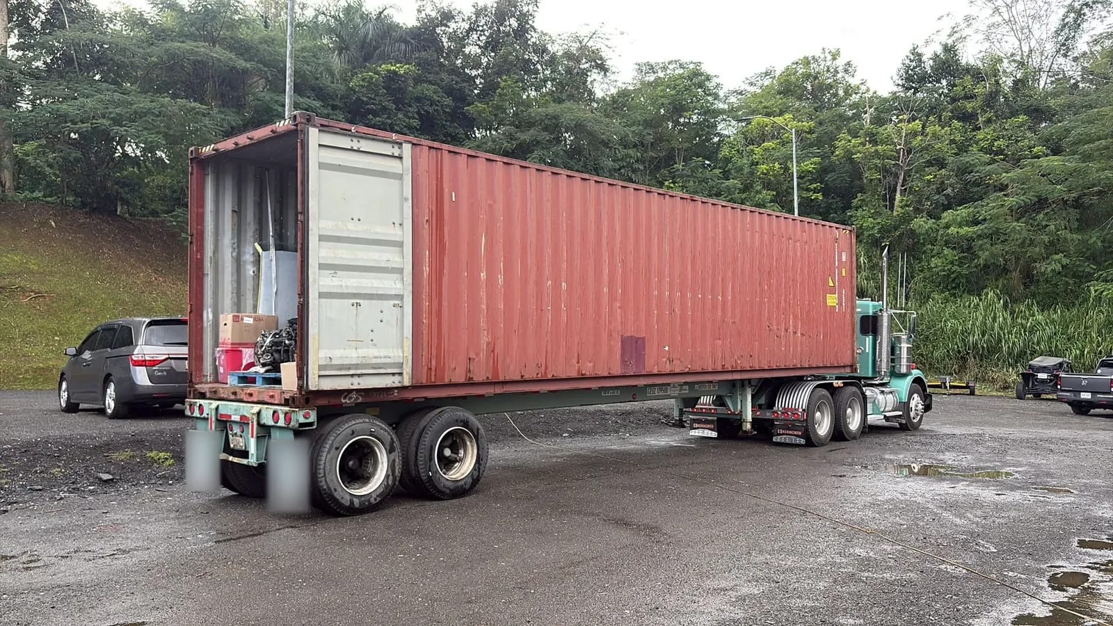 Container on a truck chassis with its rear doors open, boxes and a machine part visible inside