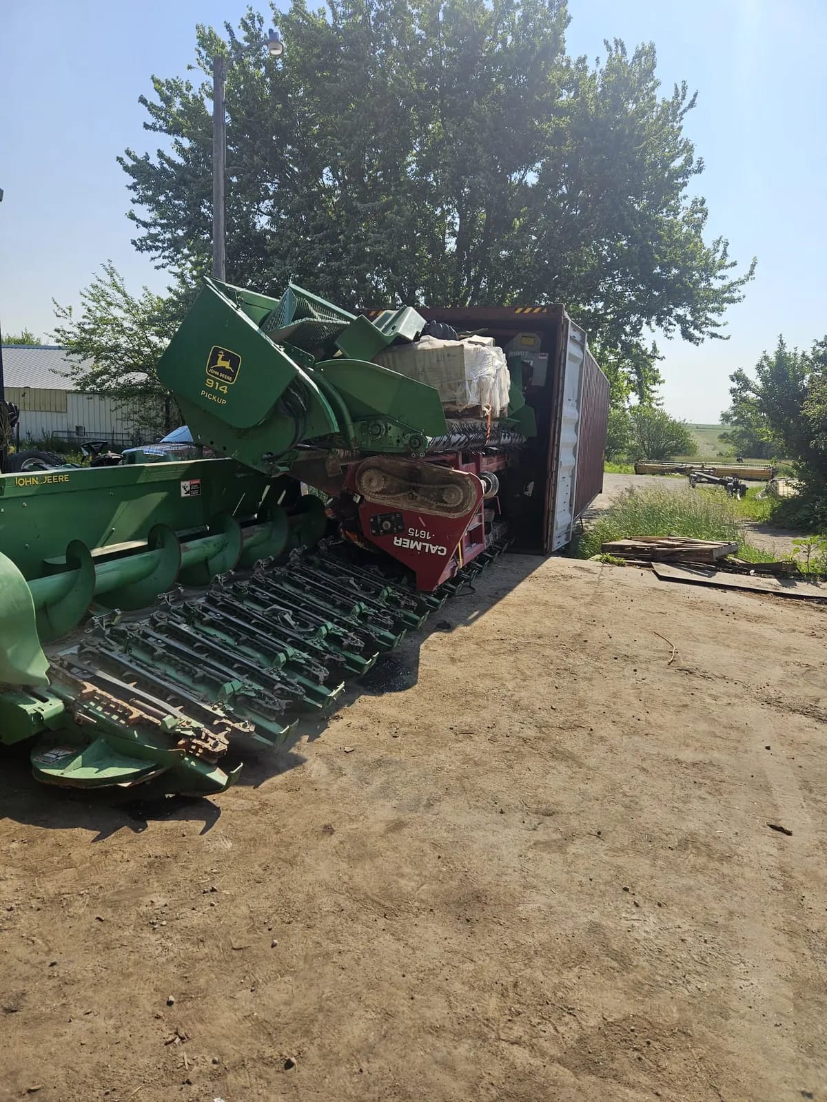 Crop headers in green and red staged beside the open end of a container with wrapped cargo inside, on a dirt yard